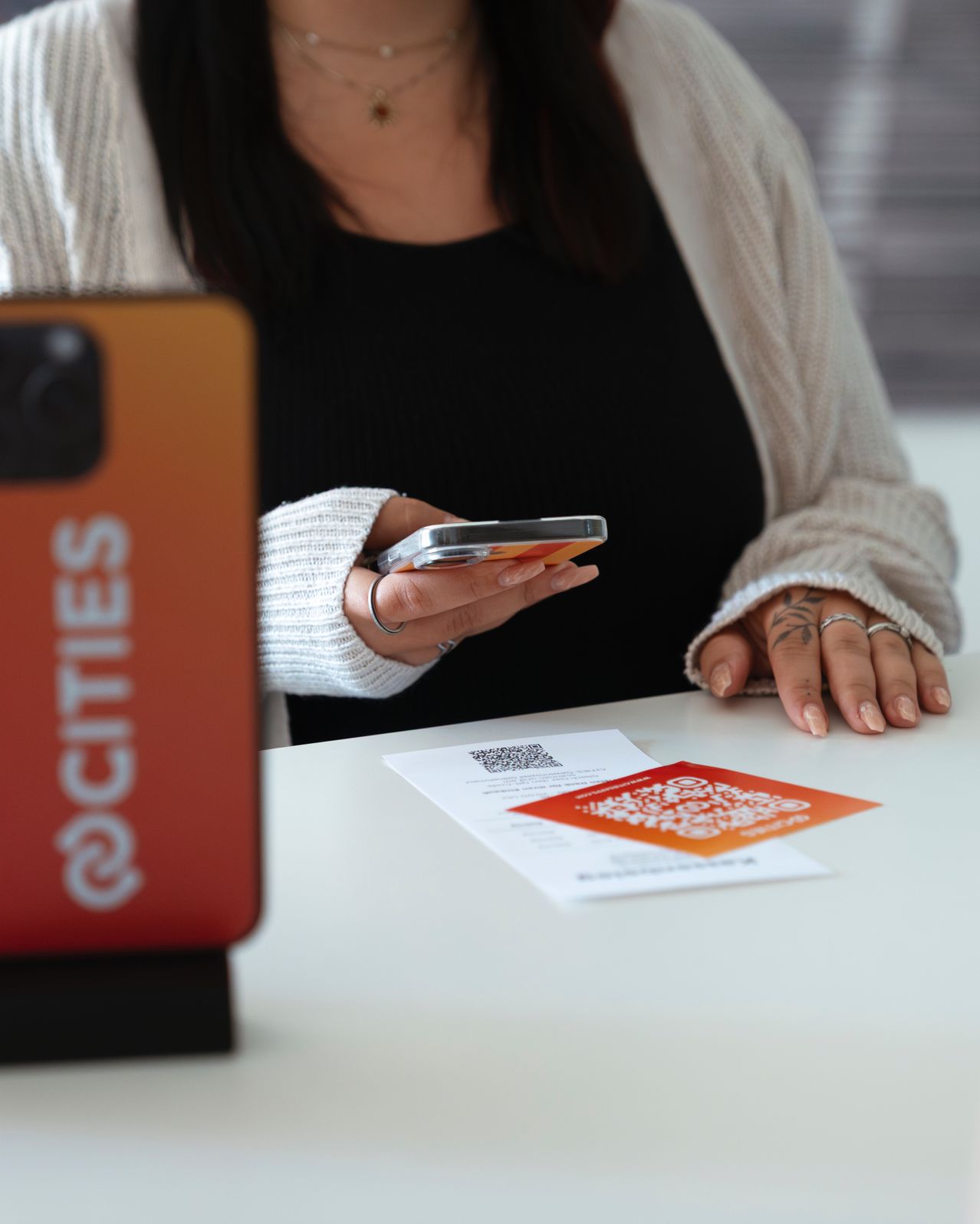 Eine Frau sitzt an einem Tisch und hält ein Smartphone und eine QR-Karte. Auf der linken Seite befindet sich ein orangefarbenes Gerät mit dem Wort 'cities'.