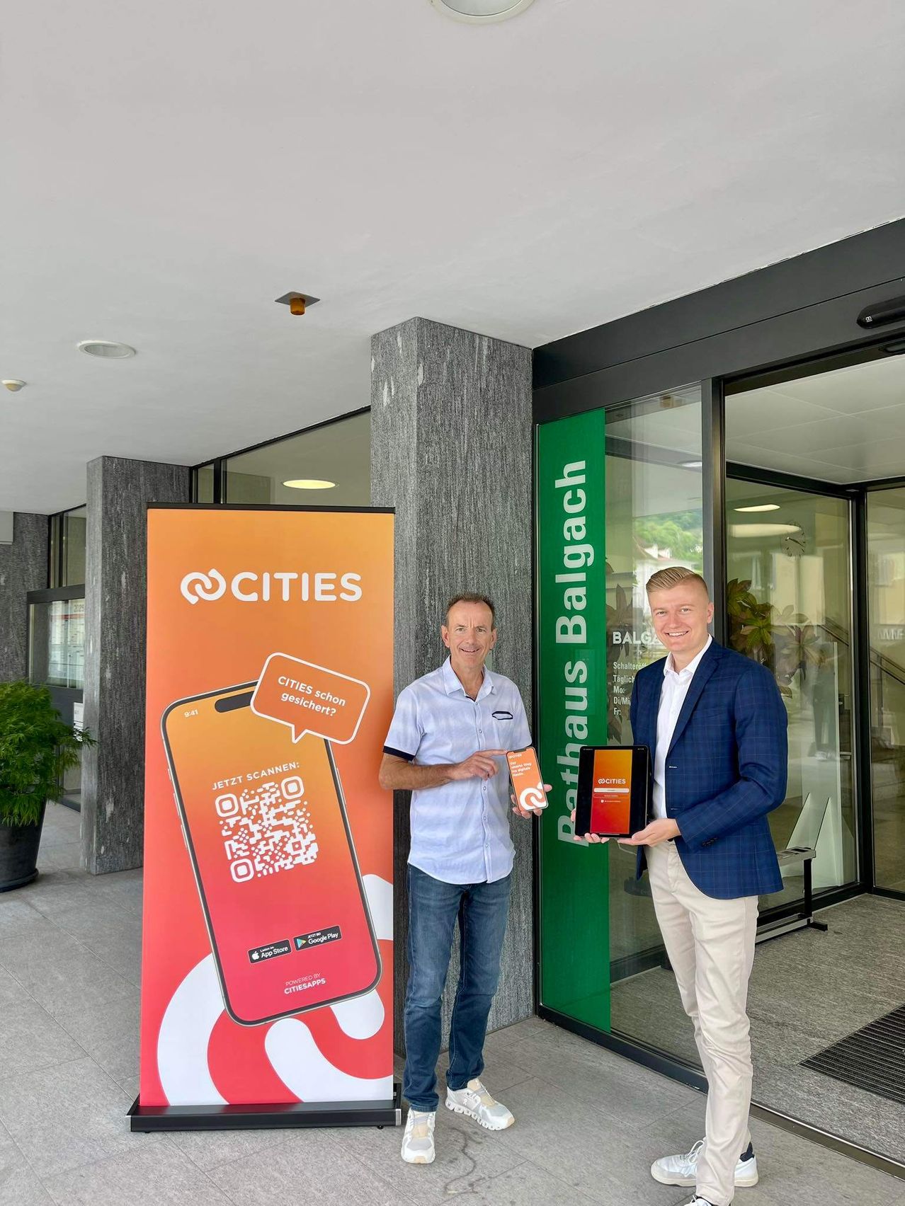 Zwei Männer stehen vor einem Gebäudeeingang, einer zeigt auf ein Smartphone mit einem QR-Code und der andere hält ein Tablet. Ein Schild hinter ihnen zeigt 'Pathhaus Balgach' und 'Cities'.