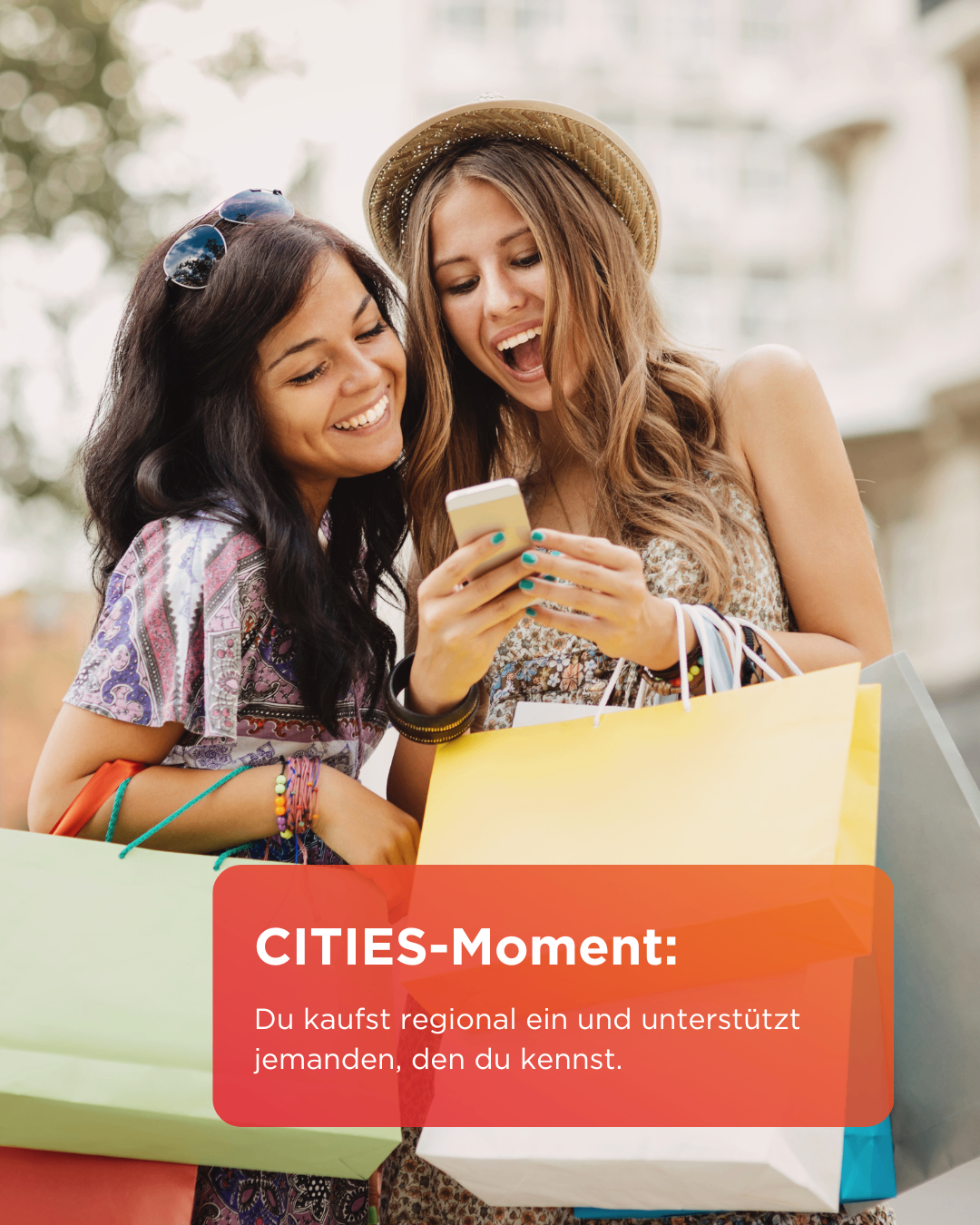 Zwei lächelnde Frauen mit Einkaufstüten betrachten ein Smartphone. Eine Frau trägt einen Hut und Sonnenbrille auf dem Kopf. Die Frau auf der rechten Seite lacht. Der Text lautet 'CITIES-Moment: Du kaufst regional ein und unterstützt jemanden, den du kennst.'