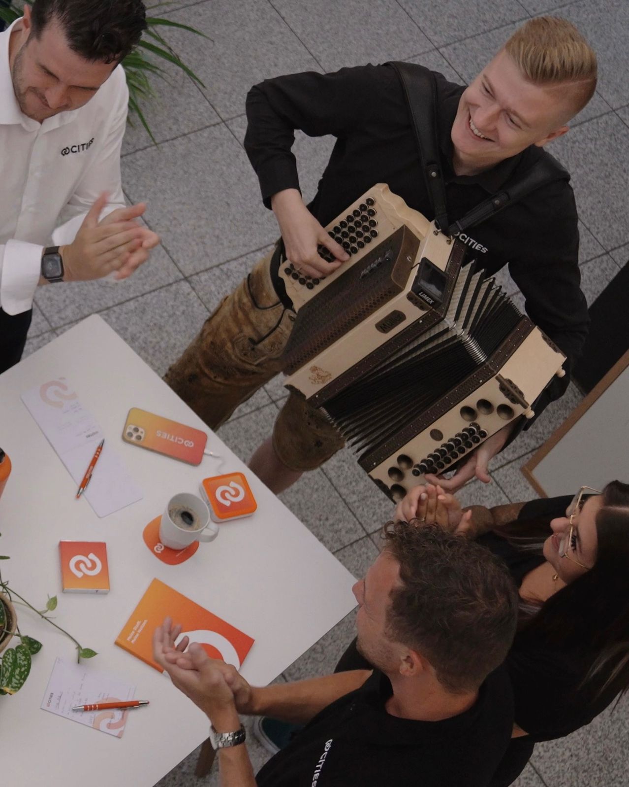 Eine Gruppe von Menschen um einen Tisch, einer spielt Akkordeon, während andere klatschen. Auf dem Tisch stehen Tassen, ein Handy und ein Stift. Ein Mann in einem weißen Hemd schaut zu.