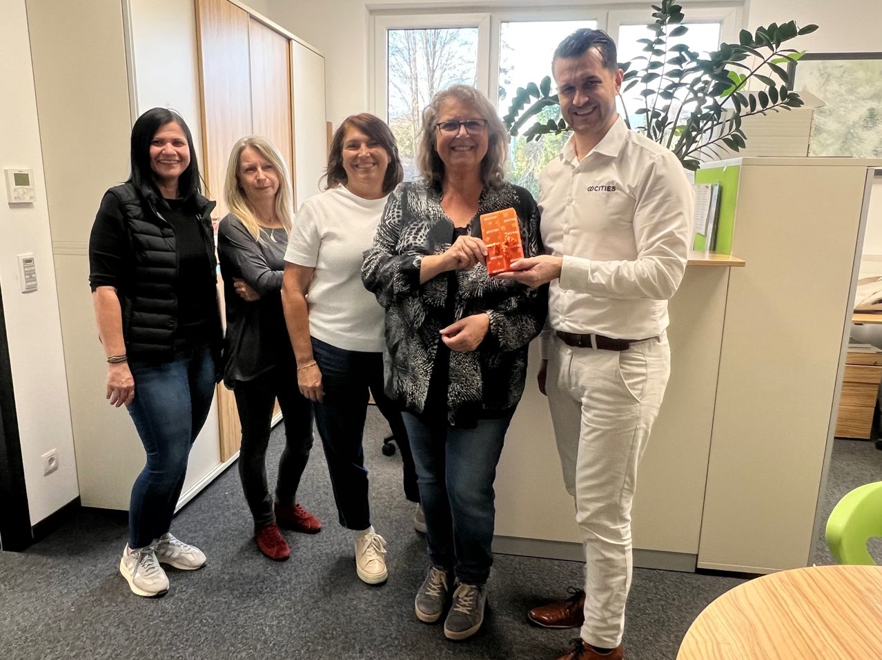 Five people stand in an office, smiling. A woman in the center holds a small orange gift box. A man on the right smiles and holds the box's ribbon. Behind them, a plant and windows with trees outside.