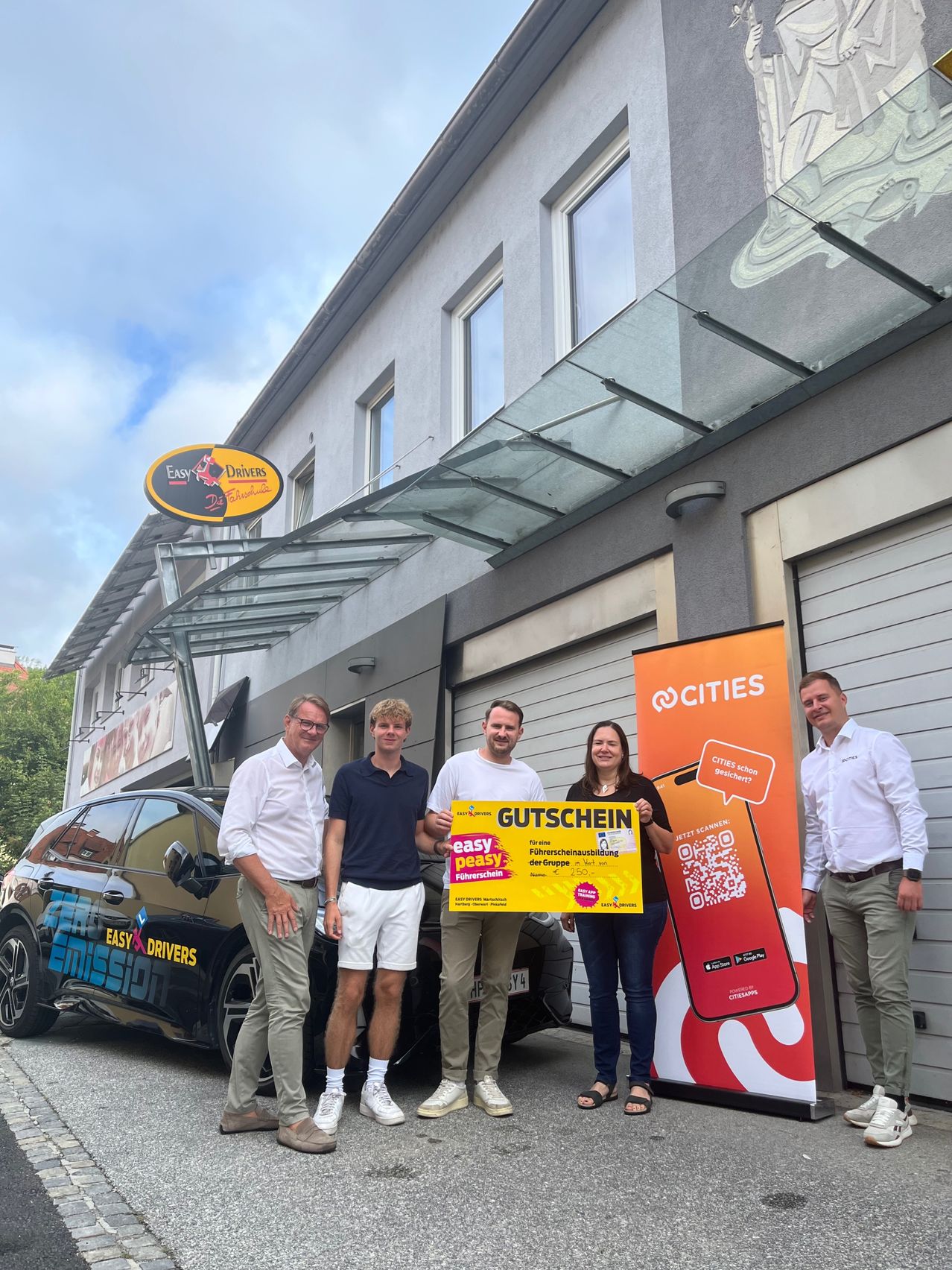 Five people stand in front of a building with a car, one holding a yellow sign that says 'easy peasy'.