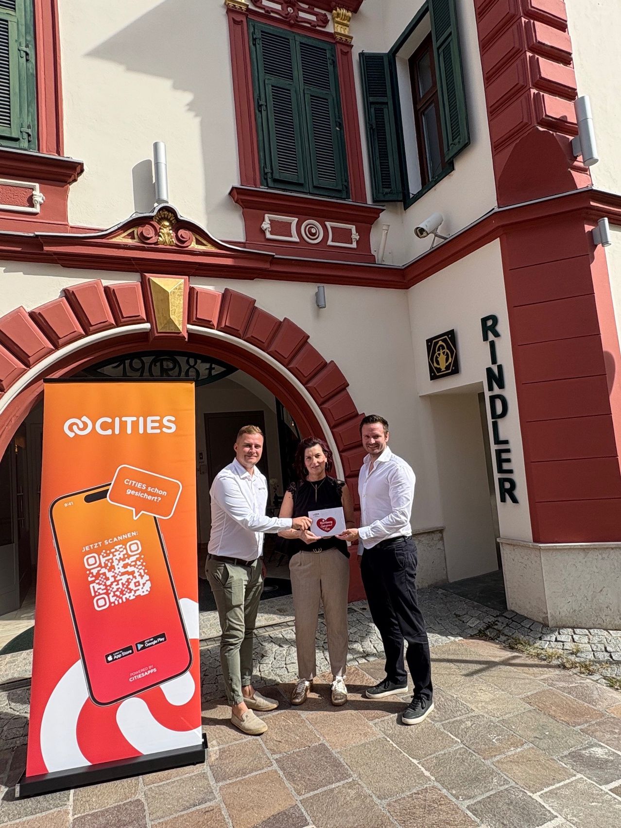 Three people stand in front of a building, two men and a woman, holding a red heart. The man on the left holds a banner with a QR code. The building has a red arch and the name Rindler on the right.