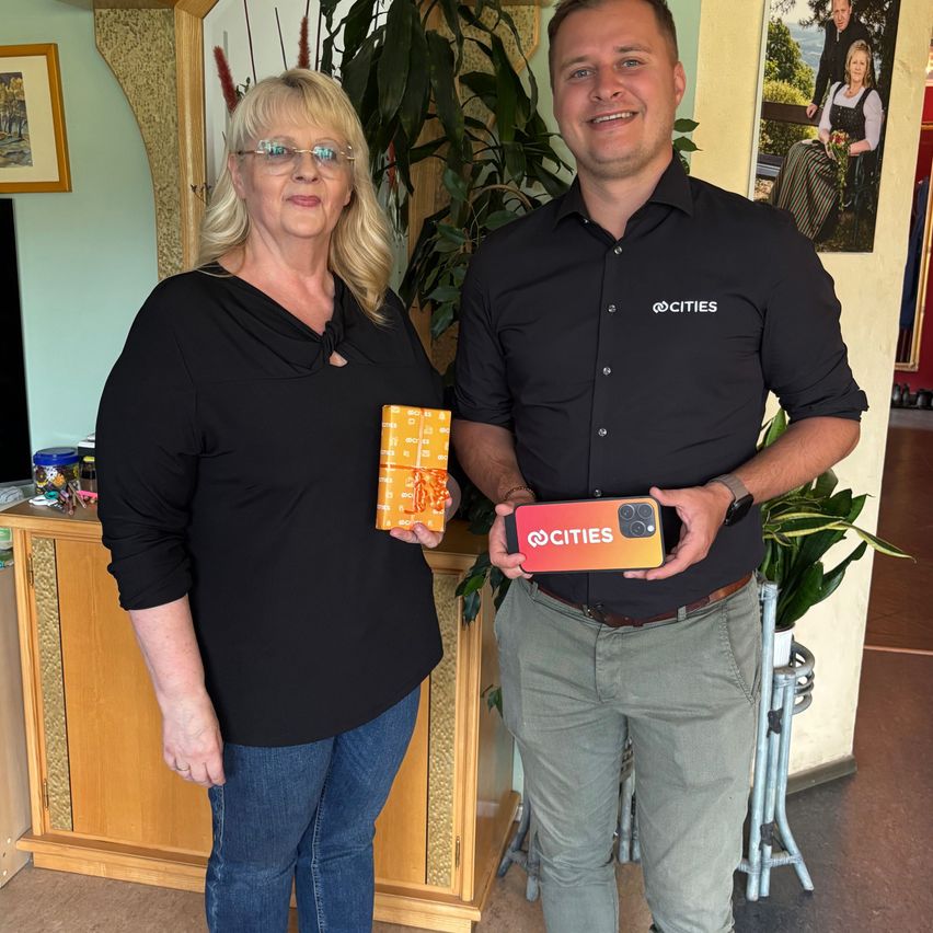 Ein Mann und eine Frau stehen lächelnd eng nebeneinander. Die Frau hält ein eingewickeltes Geschenk. Der Mann hält eine Telefonhülle mit dem Wort 'CITIES' darauf. Dahinter steht ein Holzschreibtisch mit einer Topfpflanze.