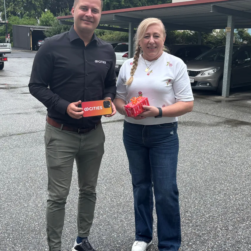 Ein Mann und eine Frau stehen in einem Parkplatz, halten Geschenke, mit einem Auto unter einem Baldachin im Hintergrund.