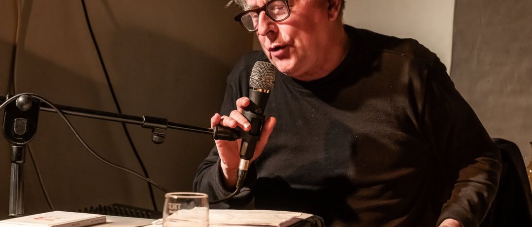 Ein älterer Mann mit Brille spricht in ein Mikrofon an einem Tisch mit Papieren und einem Glas Wasser.