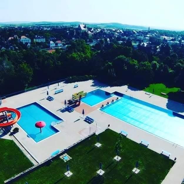 Eine Vogelperspektive eines Wasserparks mit zwei Schwimmbecken, einer Rutsche und Liegestühlen. Der Park ist von saftigem grünem Gras und Bäumen umgeben. In der Ferne gibt es Gebäude und einen klaren blauen Himmel.
