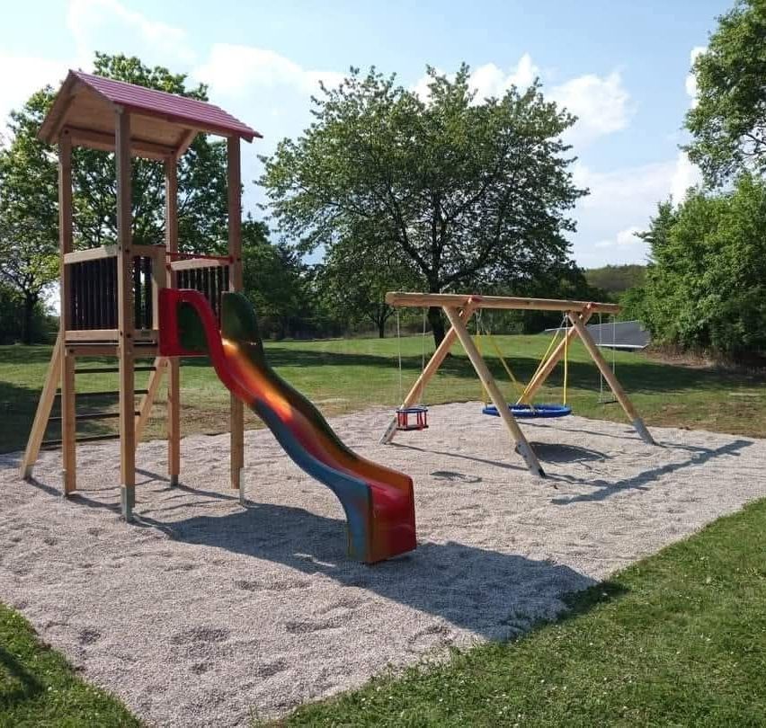 Ein Kinderspielplatz mit einer Rutsche und Schaukeln, umgeben von Gras und Bäumen an einem sonnigen Tag.