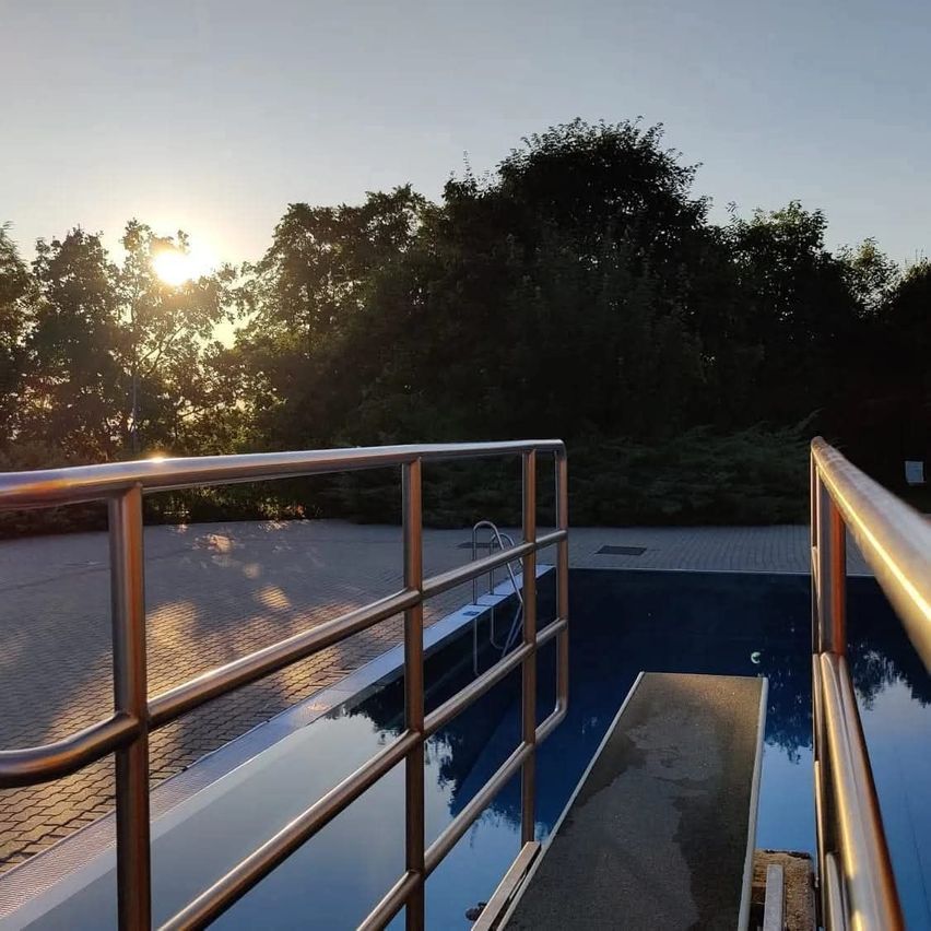 Sonnenuntergang über einem Pool, umgeben von Bäumen mit einem Metallgeländer. Die Sonne geht im Hintergrund unter.
