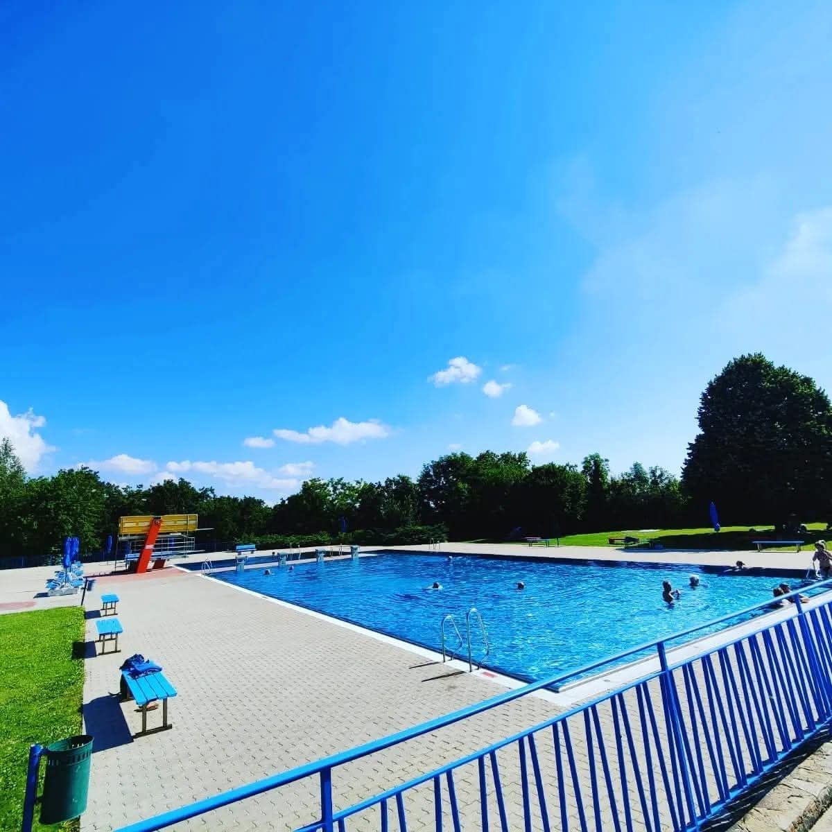 Ein Freibad mit blauen Geländern und einer blauen Rutsche an einem sonnigen Tag. Mehrere Menschen schwimmen und andere stehen an der Seite.