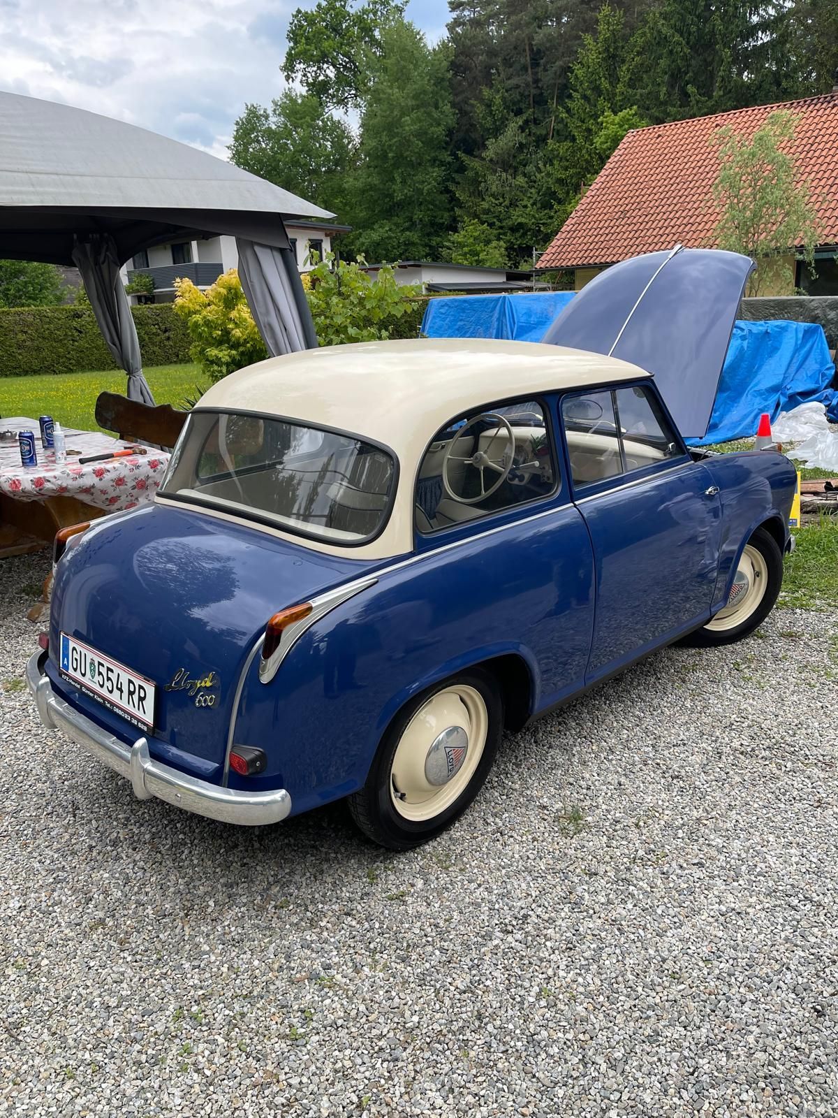 Ein blaues Oldtimer-Auto mit offenem Motorhaube, auf Kies geparkt. Dahinter ein Haus mit rotem Dach und einem Zelt im Hintergrund.