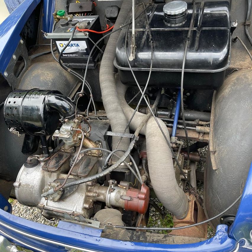 Das Innere eines Oldtimers zeigt einen freiliegenden Motor mit einem großen Auspuffrohr, einer Batterie und verschiedenen Kabeln und Drähten.