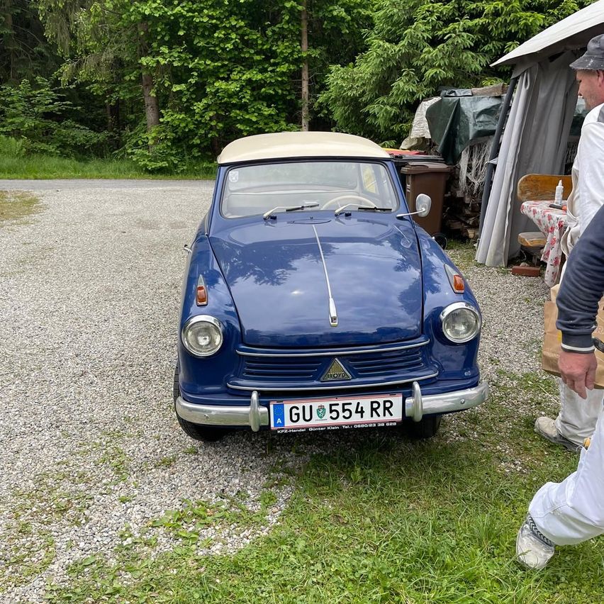 Ein altes blaues Auto mit dem Kennzeichen GU 554 RR steht auf einem Kiesweg. Zwei Männer in weißen Hosen stehen in der Nähe. Hinter dem Auto steht ein Zelt und ein Tisch mit einer Flasche.