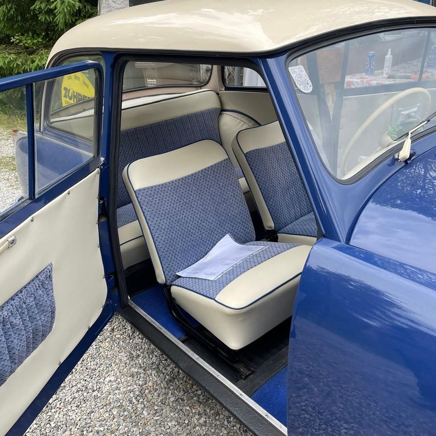 Der Innenraum eines Vintage-blauen Autos mit beigefarbenen und blauen Sitzen und einem weißen Lenkrad. Das Auto ist auf Kies geparkt.
