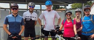 Eine Gruppe von Radfahrern in Helmen und Fahrradausrüstung posiert mit ihren Fahrrädern in einem Park. Einer hat eine Flasche am Fahrrad.