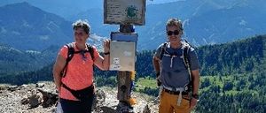 Zwei Wanderer posieren auf einem Gipfel, eine Frau in einem rosa T-Shirt und der andere Mann in einem grauen T-Shirt und orange Shorts. Sie stehen neben einem Holzkreuz und sind von felsigem Gelände und einer Gebirgslandschaft umgeben.