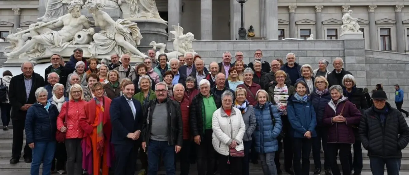 Eine Gruppe von Menschen steht auf den Stufen eines Gebäudes mit einer Statue dahinter. Sie sind in Winterkleidung gekleidet. Das Gebäude hat Säulen und Fenster.