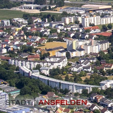 Eine Luftaufnahme einer Stadt mit vielen Gebäuden, einige mit Solarpanelen, und einer Autobahn in der Ferne. Bäume und Gras sind in der Gegend vorhanden.
