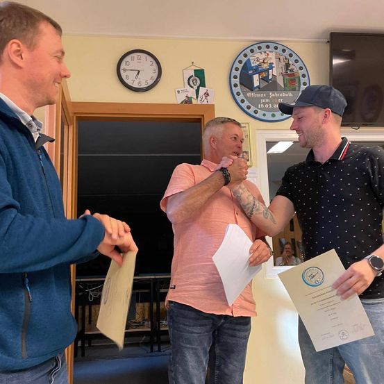 Drei Männer stehen in einem Raum und halten Urkunden in den Händen. Ein Mann in einem blauen Jacken lächelt, ein anderer in einem rosa Hemd hat eine Tätowierung und der dritte in einem schwarzen Hemd trägt eine Kappe. Eine Uhr und ein gerahmtes Bild hängen an der Wand.
