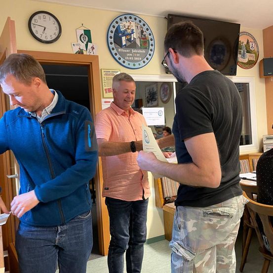 Drei Männer stehen in einem Raum, einer hält ein Zertifikat, ein anderer schaut es an und der dritte mit einer Uhr und einem Abzeichen hinter ihnen.