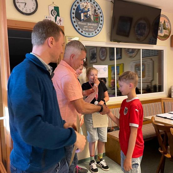 Zwei Männer überreichen einem Jungen eine Urkunde. Der Junge trägt ein rotes Shirt. Ein Mädchen steht hinter ihnen. Sie befinden sich in einem Raum mit einem großen Fenster. Es gibt einen Tisch mit Papieren darauf.