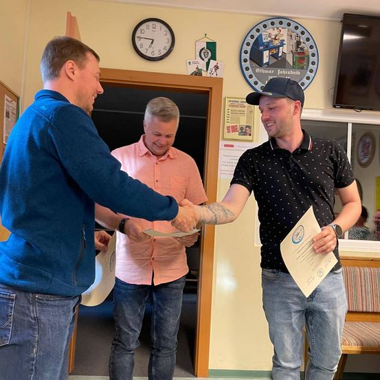 Drei Männer stehen in einem Raum, zwei schütteln sich die Hände, während der dritte ein Zertifikat hält. Sie alle lächeln. Der Raum hat eine Uhr, ein gerahmtes kreisförmiges Kunstwerk und einen Fernseher an der Wand.