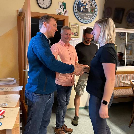 Drei Menschen stehen in einem Raum, zwei Männer und eine Frau. Sie schütteln sich die Hände. Der Mann auf der linken Seite trägt eine blaue Jacke. Die Frau auf der rechten Seite trägt ein schwarzes T-Shirt und hat eine Uhr. Der Mann in der Mitte trägt ein rosa Hemd. Hinter ihnen steht eine Uhr, eine Tafel und ein runder Bilderrahmen.