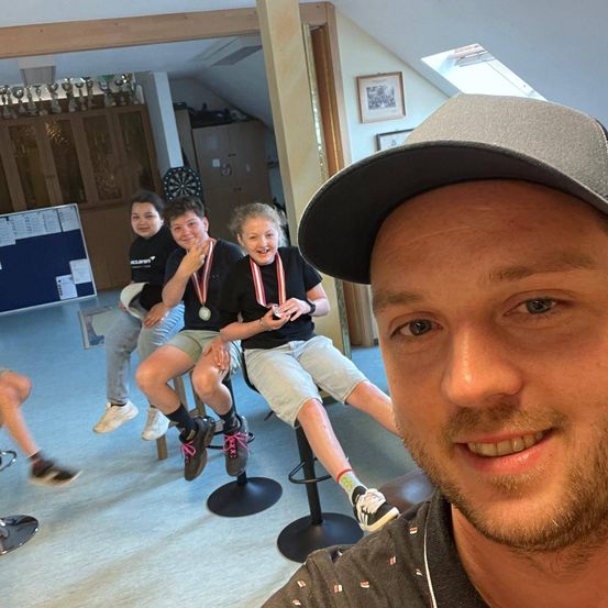 Drei Personen sitzen auf Hockern in einem Raum. Sie tragen Medaillen. Ein Mann mit einer Kappe lächelt vor ihnen.