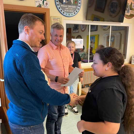 Zwei Männer und eine Frau sind in einem Raum. Ein Mann überreicht einem anderen Mann ein Papier. Eine Frau lächelt den Mann an, der das Papier erhält.