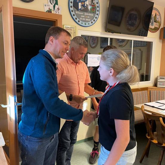 Ein Mann in einer blauen Jacke überreicht einer jungen Frau in einem schwarzen Shirt ein Zertifikat, während zwei Männer in orangefarbenen und schwarzen Shirts hinter ihnen stehen. Sie befinden sich in einem Raum mit einer Holztür, einem Schreibtisch und einem Fernseher an der Wand.