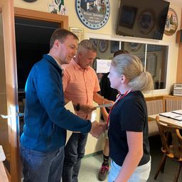 Ein Mann in einer blauen Jacke überreicht einer jungen Frau in einem schwarzen Shirt ein Zertifikat, während zwei Männer in orangefarbenen und schwarzen Shirts hinter ihnen stehen. Sie befinden sich in einem Raum mit einer Holztür, einem Schreibtisch und einem Fernseher an der Wand.