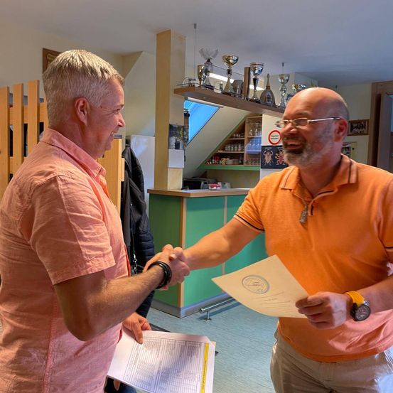 Zwei Männer in orangefarbenen Hemden schütteln sich die Hände, einer hält ein Zertifikat. Sie befinden sich in einem Raum mit ausgestellten Trophäen.