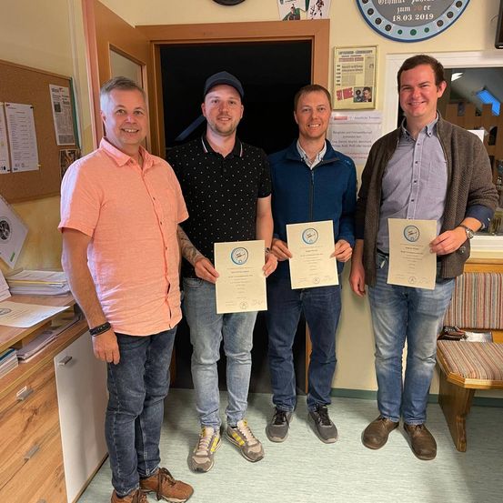 Vier Männer stehen in einem Raum, lächeln und halten Urkunden in den Händen. Einer trägt einen Hut, ein anderer eine Jacke. Hinter ihnen befindet sich eine Pinnwand mit Papieren auf einem Schreibtisch. Eine Uhr und ein gerahmtes Bild schmücken die Wand.