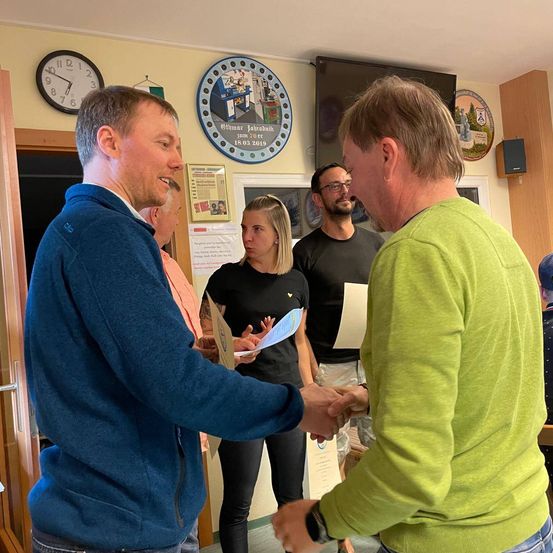 Zwei Männer schütteln sich die Hände in einem Raum. Eine Frau steht in der Nähe und hält ein Papier. Andere Personen sind im Hintergrund. Eine Uhr und ein gerahmtes Bild hängen an der Wand.