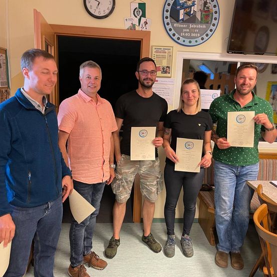 Fünf Personen stehen in einem Raum, lächeln und halten Urkunden in den Händen. Sie sind von Holzmöbeln und einer Wanduhr umgeben. Ein Monitor ist an der Wand montiert.