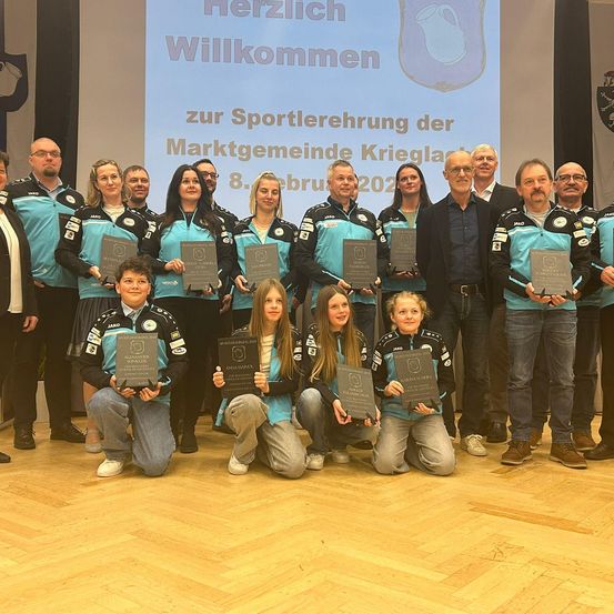 Eine Gruppe von Athleten und Trainern steht zusammen auf einer Bühne und hält Plaketten. Sie tragen passende blaue und schwarze Uniformen. Der Hintergrund zeigt eine große Leinwand mit den Worten 'Herzlich Willkommen zur Sportlehrerung der Marktgemeinde Kriegl. 8. Febr. 2022'.