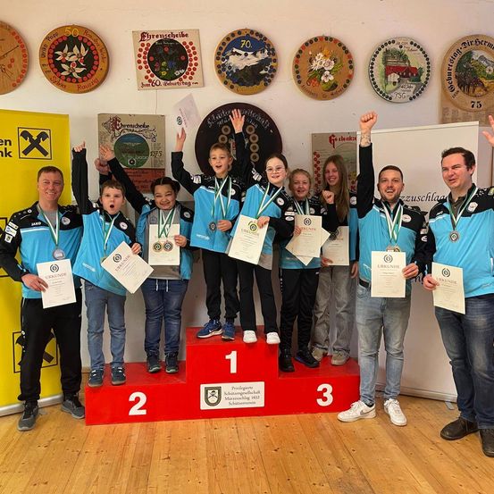 Eine Gruppe junger Athleten steht auf einem Podium mit Medaillen und Urkunden. Sie feiern ihre Leistung, und zwei Erwachsene stehen an den Seiten des Podiums. Der Raum ist mit gerahmten Tellern und Bannern dekoriert.