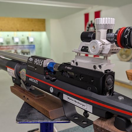 Nahaufnahme eines schwarzen Gewehrs mit rotem Streifen auf der Seite, beschriftet mit Anschutz, mit Zielfernrohr und anderen Ausrüstungsteilen.