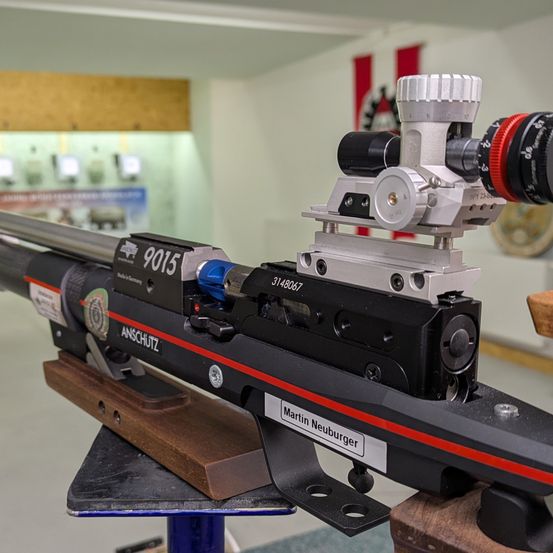 Nahaufnahme eines schwarzen Gewehrs mit rotem Streifen auf der Seite, beschriftet mit Anschutz, mit Zielfernrohr und anderen Ausrüstungsteilen.