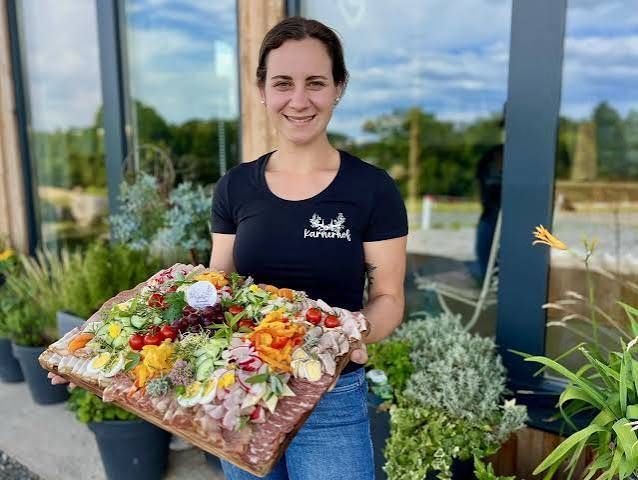Eine Frau in einem schwarzen T-Shirt hält eine große Platte mit verschiedenen Lebensmitteln, einschließlich Fleisch, Gemüse und Eiern, vor einem Garten.