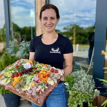 Eine Frau in einem schwarzen T-Shirt hält eine große Platte mit verschiedenen Lebensmitteln, einschließlich Fleisch, Gemüse und Eiern, vor einem Garten.
