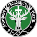 Ein kreisförmiges Emblem mit einem weißen Hirsch mit Geweih, einem Kreuz auf seiner Stirn und einem grünen Hintergrund mit Text darum herum.