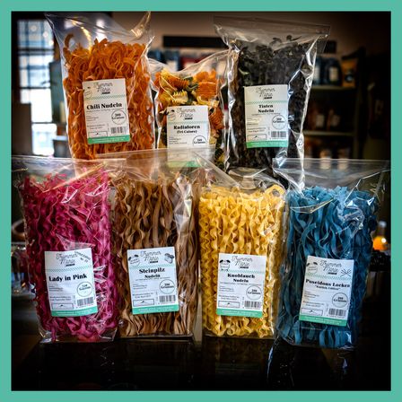 Verschiedene Packungen bunter Nudelart sind auf einem Regal ausgestellt. Beinhaltet Chili Nudeln, Radiaforen, Lady in Pink, Steinpilz Nudeln, Knoblauch Nudeln und Poseidons Locken. Jede Packung hat ein Etikett mit Name und Gewicht.