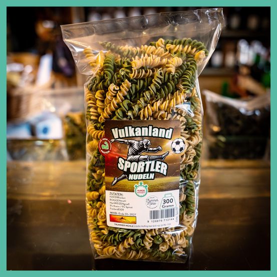 Ein Plastikbeutel mit Sportler-Nudeln und einem braunen Etikett, das einen Fußballspieler beim Balltreten zeigt. Die Nudeln sind grün und gelb.