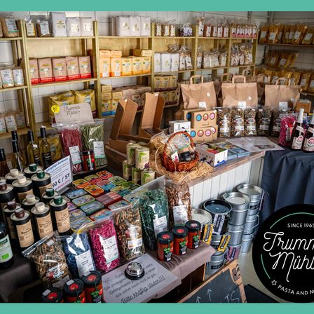 Ein Geschäft gefüllt mit verschiedenen Produkten wie Pasta, Getreide, Flaschen und Konserven auf Regalen und Tischen, mit einem Schild, das 'Seit 1965' steht.