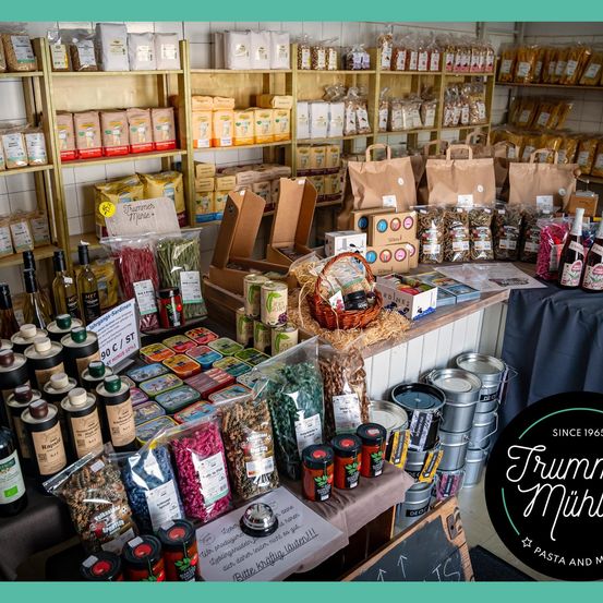 Ein Geschäft gefüllt mit verschiedenen Produkten wie Pasta, Getreide, Flaschen und Konserven auf Regalen und Tischen, mit einem Schild, das 'Seit 1965' steht.