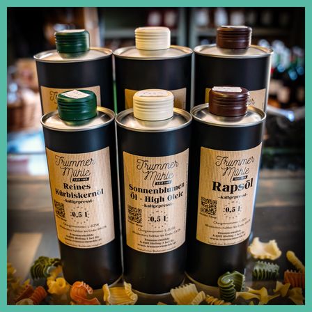 Vier Flaschen Öl in einer Reihe, beschriftet mit 'Trummer Mühle'. Jede Flasche hat einen grünen Deckel und ein Etikett mit Text und Barcode. Der Hintergrund zeigt einen Tisch mit Nudeln und einem verschwommenen Laden.