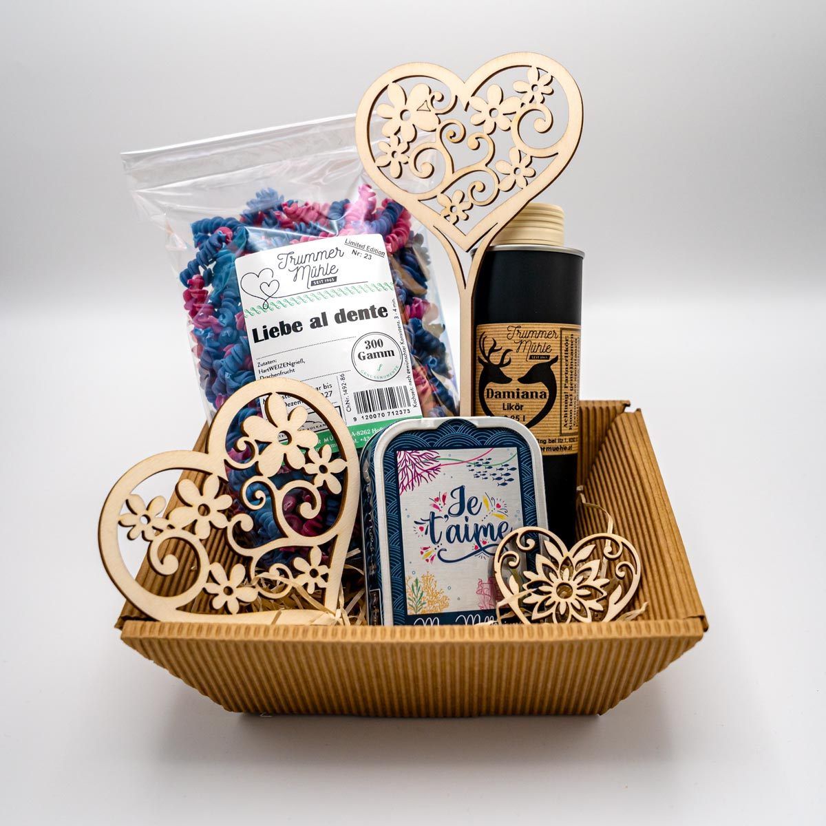 Ein herzförmiges Geschenkkörbchen mit einem Blumendesign, bestehend aus einem Beutel mit farbigen Nudeln, einer Flasche Likör, einer Karte und einer kleinen Blechdose, alles verpackt in einem braunen Papierbeutel.