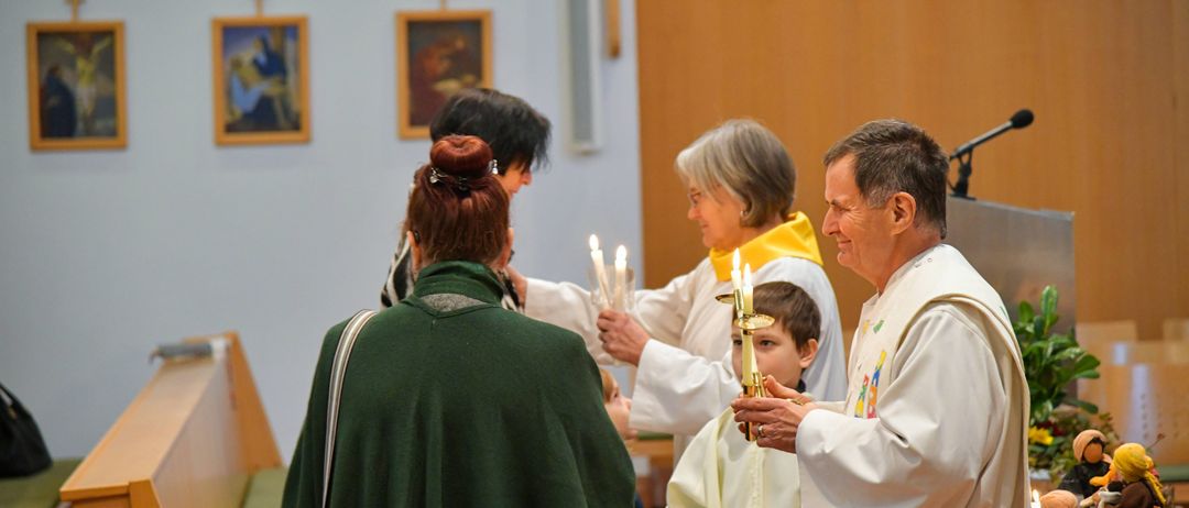 Ein Priester hält Kerzen für einen Jungen und eine Frau während einer religiösen Zeremonie. Eine weitere Frau steht hinter ihnen.