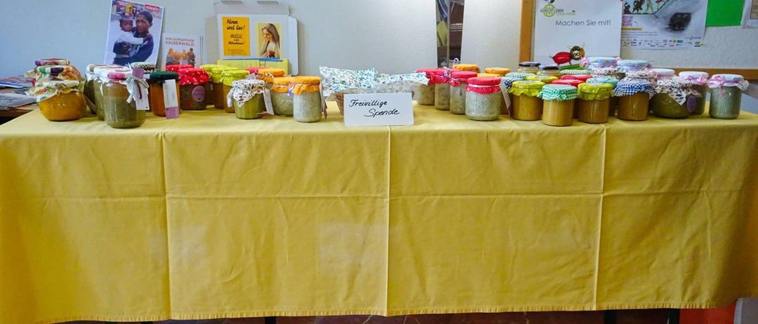 Ein Tisch mit gelber Tischdecke ist mit Gläsern verschiedener Größen und Farben bedeckt, die als 'Frohliche Spende' beschriftet sind. Hinter dem Tisch befinden sich auch Bücher und ein Plakat an der Wand.